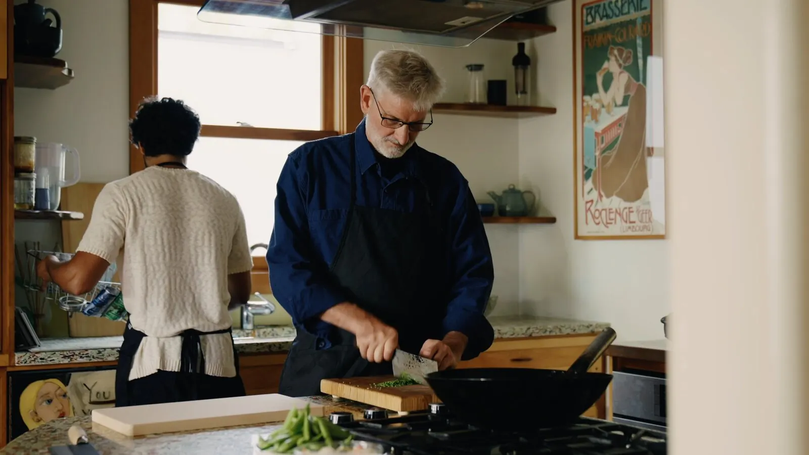 A home cook chops chives with a cleaver at the stove while Akshay Prabhu works at the counter behind him
